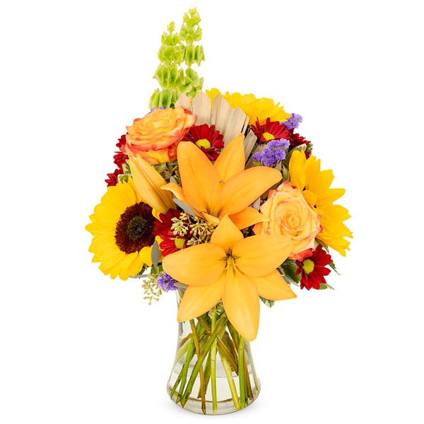 Full bouquet of lilies, roses, sunflowers, and daisies, in bright fall colors of orange, yellow, and red in a clear glass vase.