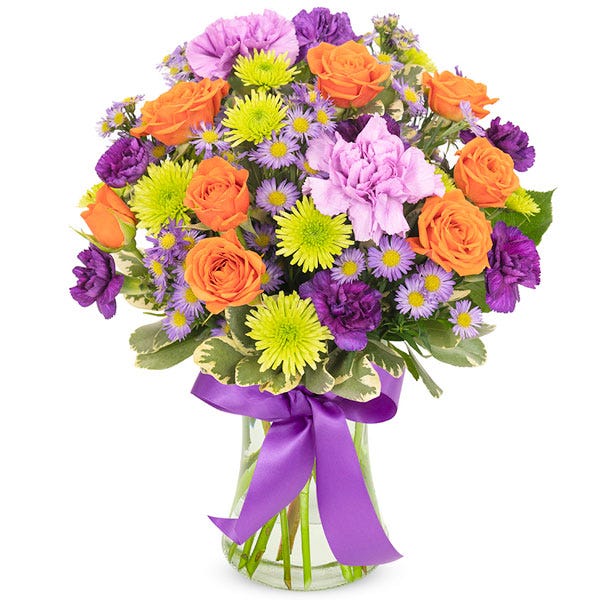 Spray roses and carnations in shades of orange, purple, and green arranged in a clear glass vase with purple ribbon.
