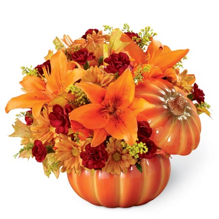 A bouquet of Orange Asiatic Lilies, Burgundy Mini Carnations, Butterscotch Daisies, and Lush Greens on a Ceramic Pumpkin Container With Lid