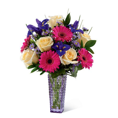Yellow roses, hot pink gerbera daisies and purple iris in a purple vase