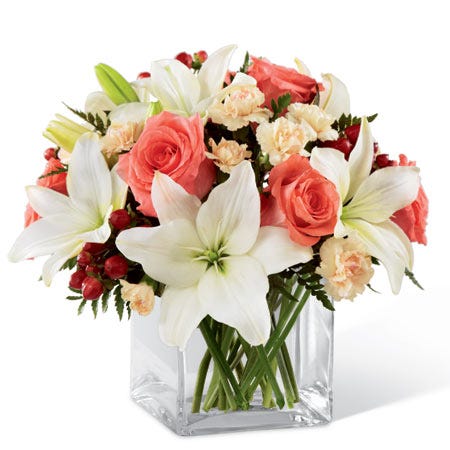 Coral roses, white Asiatic lilies, peach mini carnations, and red hypericum berries in a clear glass cube vase