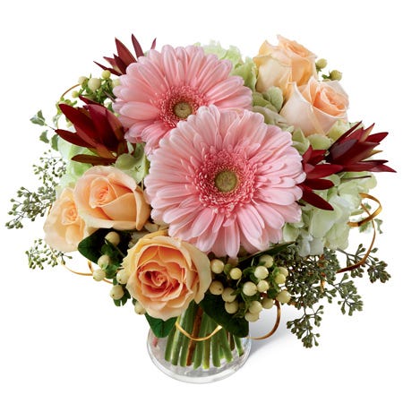Pink gerbera daisies, peach roses, green hydrangea, peach hypericum berries, & lush greens in a clear glass vase
