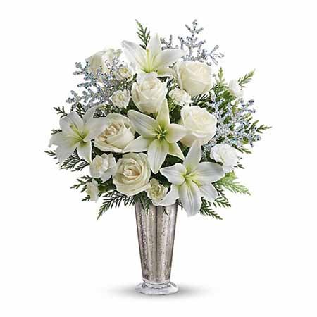 A bouquet of White Roses, White Lilies, White Mini Carnations, Flat Cedar Branches, and Silver Snowflakes in a Silvery Vase