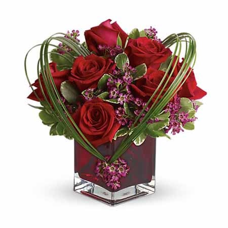 A glass scarlet colored vase including red roses, purple waxflower variegated pittosporum and bear grass