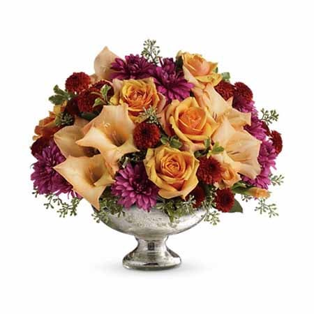 A Centerpiece flower arrangement including  Lush Greens, Orange Roses, Gladioli, Lavender Cushion Spray Mums, Orange Lilies in a Mercury Glass Vase with 1 Orange Candle