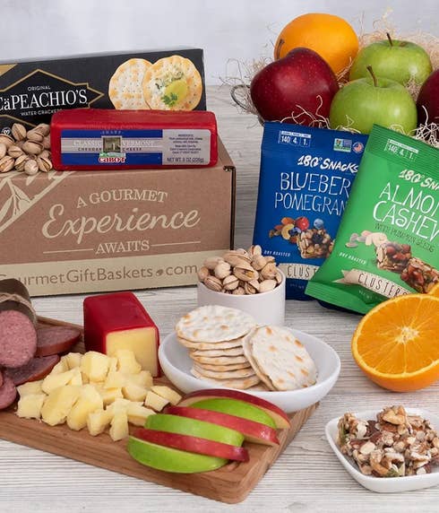 Gourmet gift box with sharp Vermont cheddar cheese, water crackers, summer sausage, pistachios, nut clusters, red and green apples, an orange half, and snack packs of blueberry pomegranate and almond cashew clusters arranged on a table.