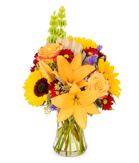 Full bouquet of lilies, roses, sunflowers, and daisies, in bright fall colors of orange, yellow, and red in a clear glass vase.