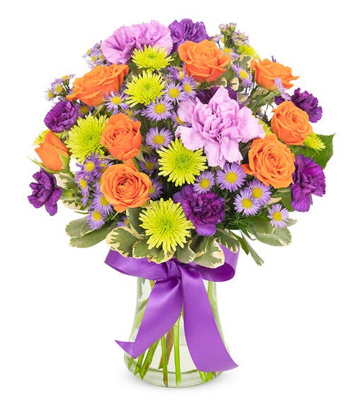 Spray roses and carnations in shades of orange, purple, and green arranged in a clear glass vase with purple ribbon.