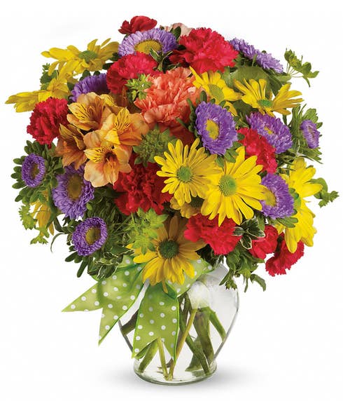 An assortment of yellow daisies, red carnations, purple matsumoto asters, orange alstroemeria, and green bupleurum in a clear glass vase with a green and white dotted ribbon bow.