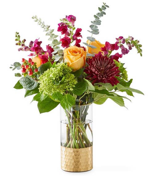 Peach Roses, Green Mini Hydrangea, Purple Snapdragons, red Disbud Poms, Gray & Green Eucalyptus, and floral greens in a half gold glass vase against a white background