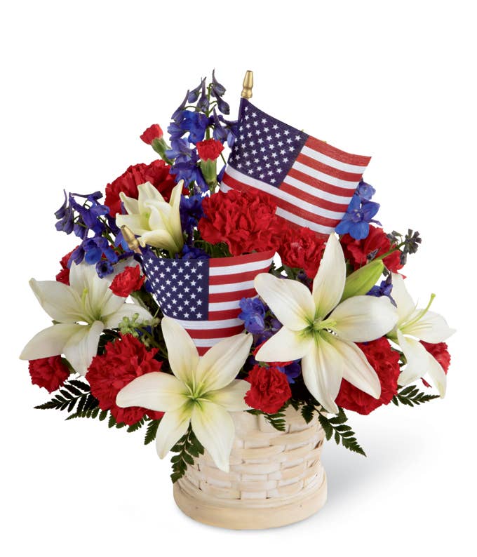 A Bouquet of Red Carnations, Red Mini Carnations, Volkenfrieden Delphinium, and White Asiatic Lilies in a Whitewash Basket with Small American Flag Inserts and Message Card