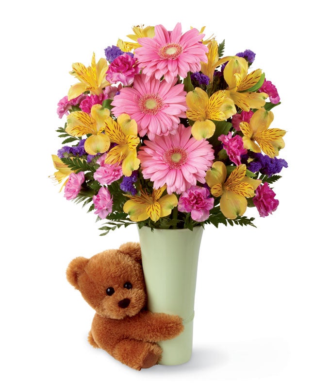 A bouquet of pink gerbera daisies, yellow Peruvian lilies, fuchsia mini carnations, and purple statice on a floral vase with a brown teddy bear hugging it