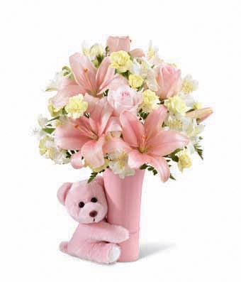 A bouquet with a combination of pastel pink roses, pink Asiatic lilies, yellow mini carnations, white Peruvian lilies and lush greens on a pink vase with a teddy bear hugging it