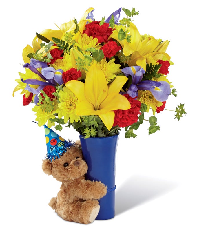 A bouquet of yellow Asiatic lilies, red roses, red carnations, yellow chrysanthemums, crimson mini carnations, blue iris and lush greens on a blue ceramic vase with a bear hugging it