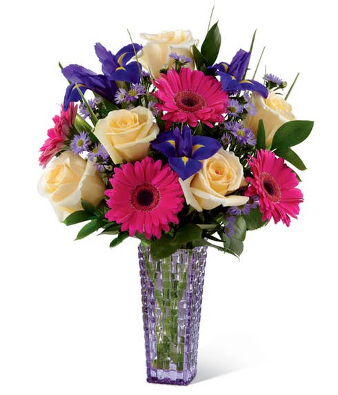 Yellow roses, hot pink gerbera daisies and purple iris in a purple vase