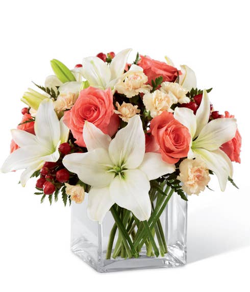 Coral roses, white Asiatic lilies, peach mini carnations, and red hypericum berries in a clear glass cube vase