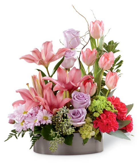 Lavender roses, pink tulips, fuchsia carnations, pink Asiatic lilies, lavender daisies, green mini hydrangea and an assortment of greens with curly willow tips in a metal oval container