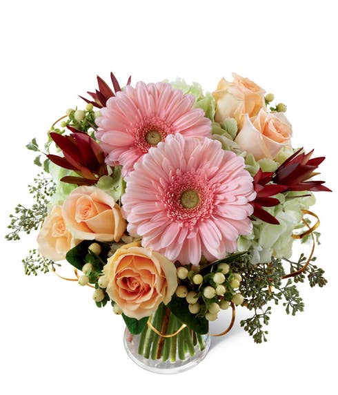Pink gerbera daisies, peach roses, green hydrangea, peach hypericum berries, & lush greens in a clear glass vase