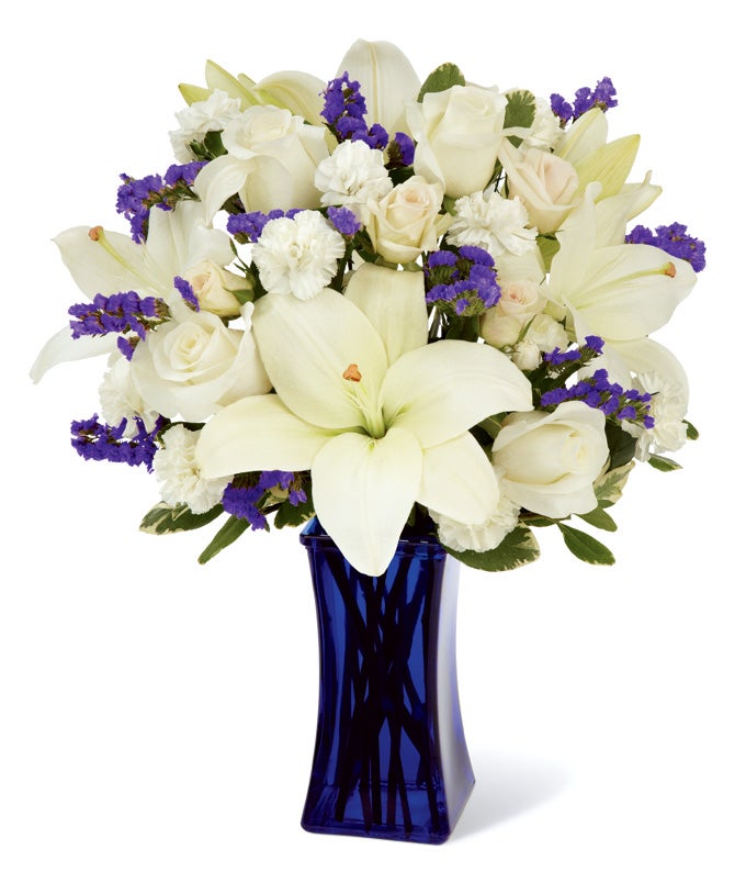 A bouquet of White Asiatic Lilies, Pale Ivory Roses, Milky Mini Carnations, Blue Statice, Cream Spray Rose and Lush Greens in a Dark Blue Glass Vase