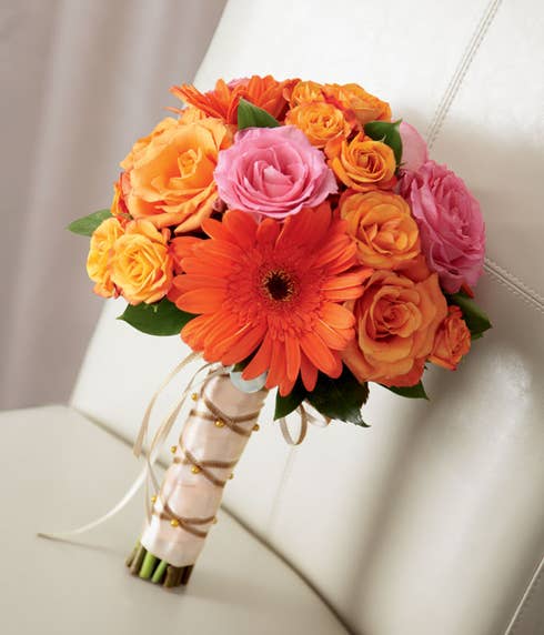 Tied bouquet of orange roses, spray roses, and gerbera daisies arranged with fuchsia roses all tied together at the stems with peach satin ribbon