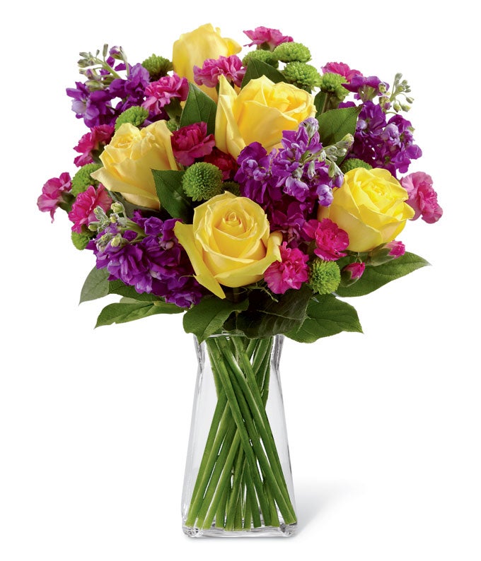 A Bouquet of Yellow Roses, Purple Stock, Green Button Poms, Fuchsia Mini Carnations, and Lush Greens in a Square Glass Vase