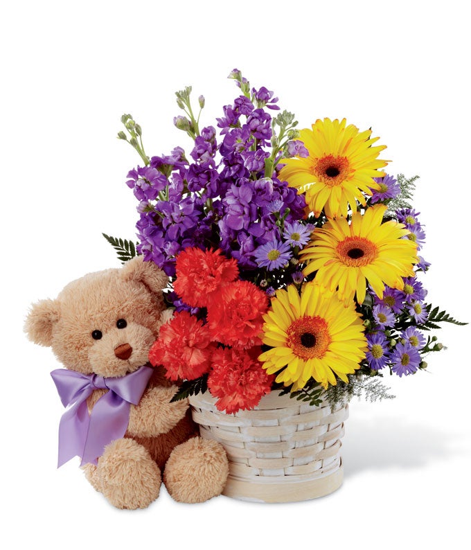 A boquet of Dark Purple Stock, Bright Orange carnations, Yellow Gerbera Daisies, Indigo Monte Casino Asters and Lush Greens on a Whitewash Woven Basket Container and a 10 Inch Plush Bear