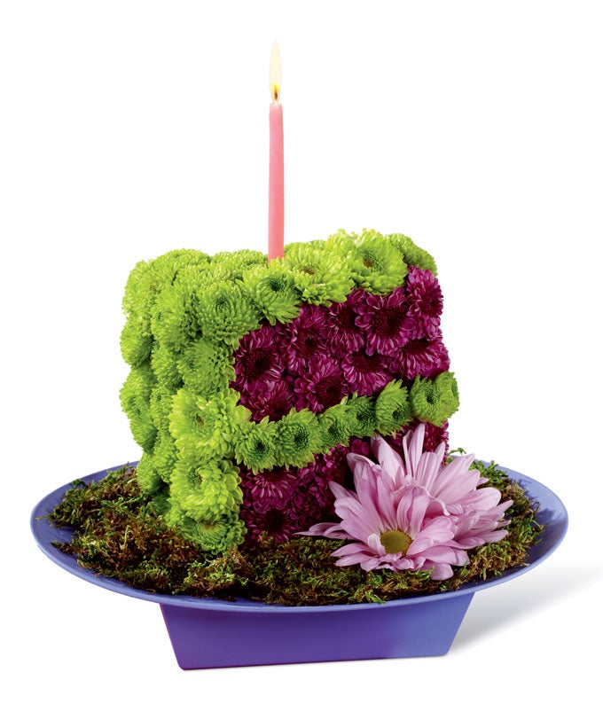A bouquet of Purple Button Poms, Green Button Poms, Sheet Moss, and Purple Daisies formed as 1 slice of cake with pink candle on an oval bowl
