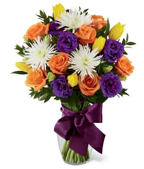Orange roses, yellow tulips, white spider chrysanthemums, purple double lisianthus and myrtle in a clear glass vase with a mauve satin ribbon