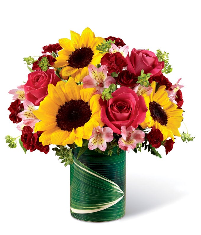 A bouquet of sunflowers, pink roses, pink alstroemeria, burgundy mini carnations and lush greens on an illusion faux leaf vase
