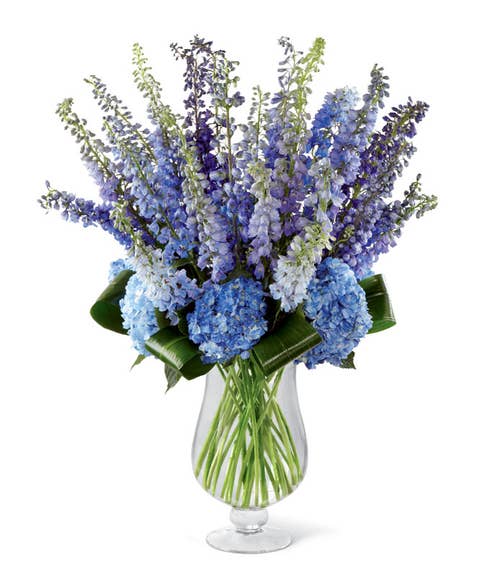 Blue delphinium, purple delphinium, dark blue hydrangea, and aspidistra leaves in a footed vase