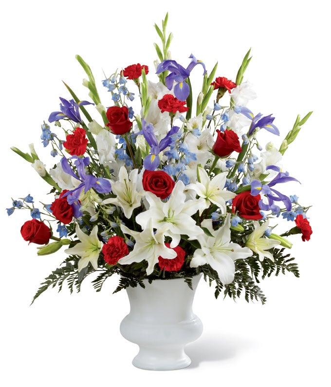 A Bouquet of  Red Roses, Red Carnations, White Gladiolus, Light Blue Delphinium, Blue Iris, and White Lilies in a White Plastic Urn