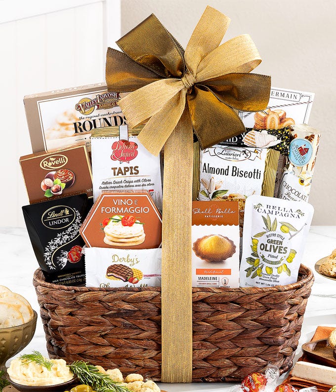 Wicker gift basket with a brown and cream bow, filled with gourmet snacks including cheese spread, chocolate chip cookies, lemon cake, hummus, crackers, dark chocolate sea salt caramels, and assorted sweet and savory treats