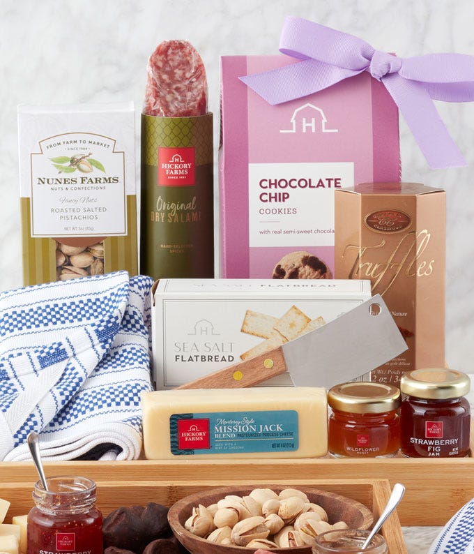 Mother's Day gift basket on a bamboo tray with salami, cheese, pistachios, truffles, cookies, fig jam, honey, a cleaver, and tea towel, ideal for gourmet-loving moms.
