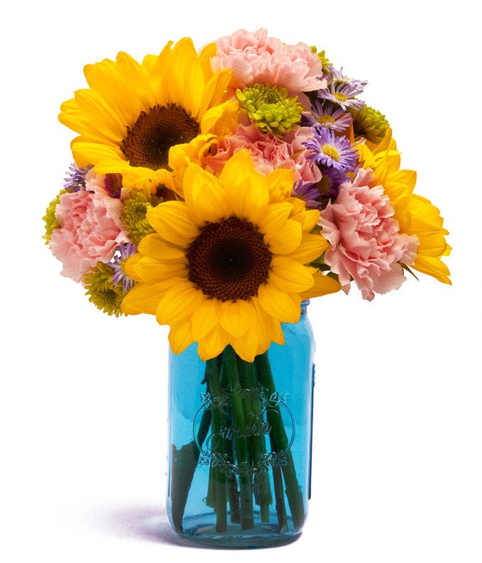 A Bouquet of Sunflowers, Pink Carnations, Green Poms and Purple Daisy Poms in a Blue Mason Jar Vase