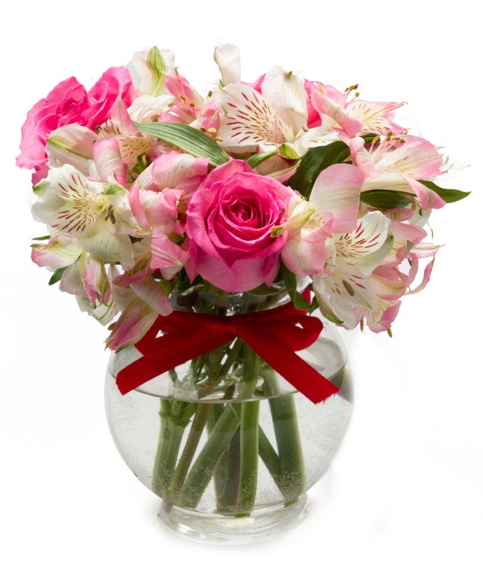 A bouquet of flowers including Pink Roses, White Alstroemeria, and Fresh Greens on Tiny Glass Vase with Colored Ribbon Decoration