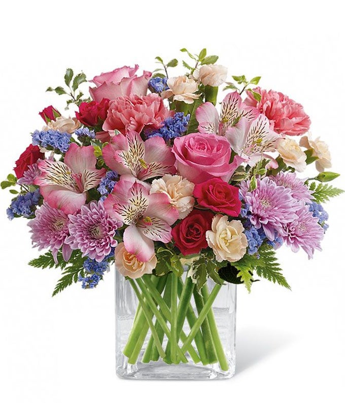 A Bouquet of Pink and Peach Mini Carnations, Light-Pink Alstroemeria, Roseate Roses only Included in Deluxe & Premium Options, Pitta Negra, Leatherleafs, Violet Sinuata Statice, and Lavender Cushion Spray Mums in a Clear Cube Vase