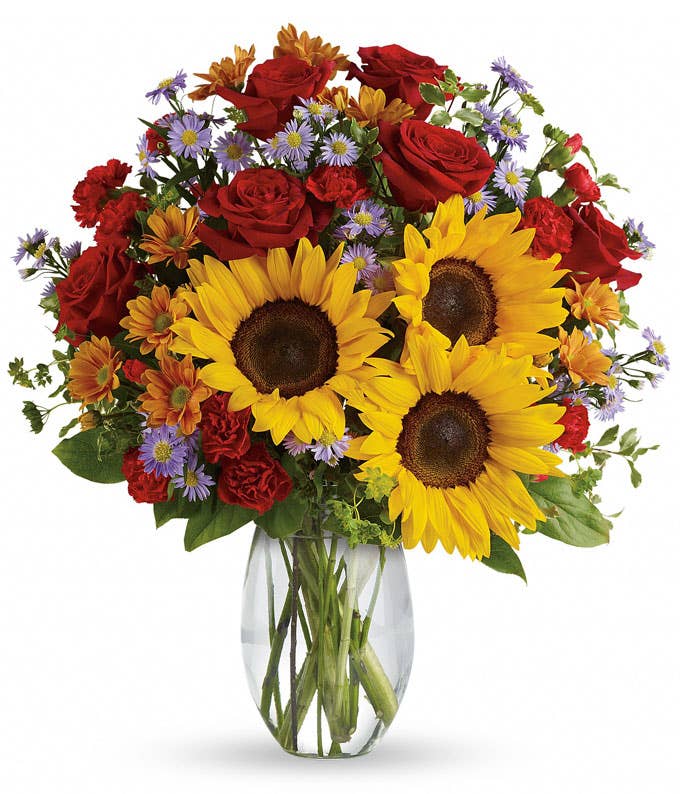 A Bouquet of  Large Sunflowers, Red Roses, Mini Red Carnations, Bronze Daisies, Lavender Monte Casino Asters, Bupleurum and Salal & Pitta Negra in a Clear Glass Vase