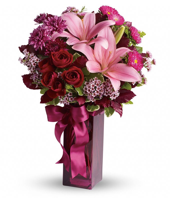 A Bouquet of Red Spray Roses, Pink Lilies Included in Deluxe and Premium Sizes Arrangement Only, Blush Carnations in Regular Size  Arrangement Only, Hot-Pink Matsumoto Asters, and Lavender Waxflowers in a Plum Colored Vase