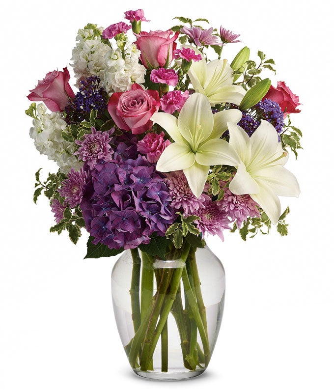 A Bouquet of Purple Hydrangea, Pink Roses, White Asiatic Lilies, Mini Pink Carnations, White Stock, Lavender Cushion Spray Chrysanthemums, Purple Seafoam Statice, and Negra Pitta in a Clear Glass Vase