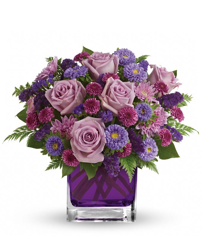 A Bouquet of  Lavender Roses, Lilac Matsumoto Asters, Purple Button Spray Chrysanthemums, Light-Purple Cushion Spray Chrysanthemums, Violet Sinuata Statice, Leatherleaf Fern, and Lemon Leaf in a Colorful Cube Glass Vase