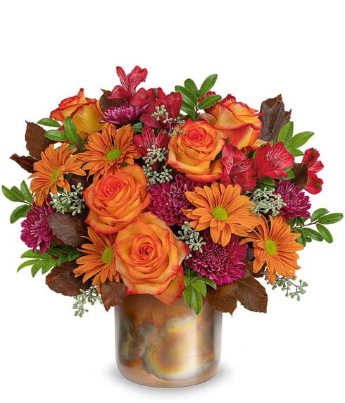 Orange roses, red altromeria. purple cushion chrysanthemums, bronze daisy spray chrysanthemums, brown copper beech, seeded eucalyptus, huckleberry in a bronze cylinder vase with patina against a white background