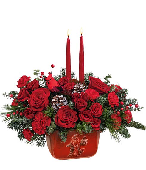 Red roses & carnations among noble fir and white pine with pinecones, berries, and cardinal picks, all within aa red ceramic bowl with a cardinal detail. Two red taper candles are placed in the floral arrangement. 