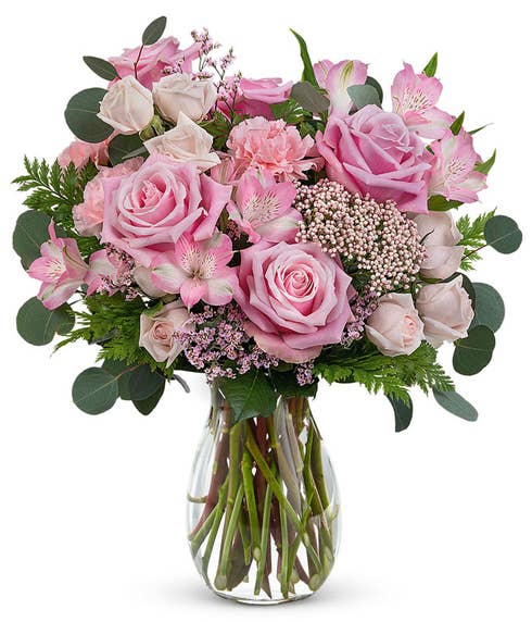 A Mother's Day arrangement featuring light pink spray roses, pink alstroemeria, carnations, limonium, rice flower, and floral greenery in a clear jordan vase, with a space for a personalized gift message.