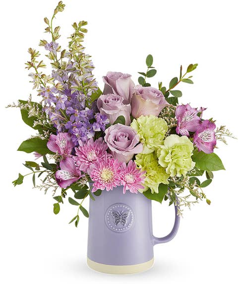 A Mother's Day arrangement featuring lavender roses, purple alstroemeria, green carnations, lavender larkspur, cushion chrysanthemums, and floral greenery, arranged in a keepsake lavender butterfly pitcher, with space for a personalized gif