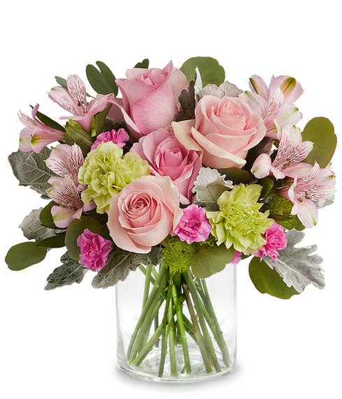 A Mother's Day arrangement featuring pink roses, light pink alstroemeria, green carnations, cushion spray chrysanthemums, and floral greens, arranged in a short clear cylinder vase, with space for a personalized gift message.