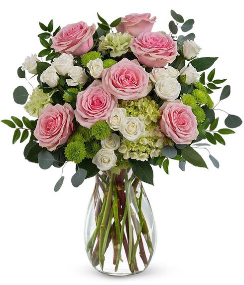 A Mother's Day arrangement featuring green hydrangea, pink roses, white spray roses, green carnations, green button chrysanthemums, and floral greenery, arranged in a clear glass jordan vase, with space for a personalized gift mes