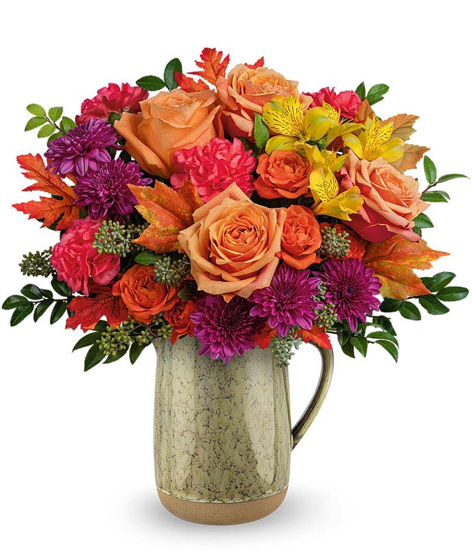 A fall floral arrangement featuring peach roses, orange spray roses, yellow alstroemeria, orange carnations, and purple cushion spray mums, accented with seasonal greenery and autumn leaves, elegantly arranged in a rustic ceramic pitcher.