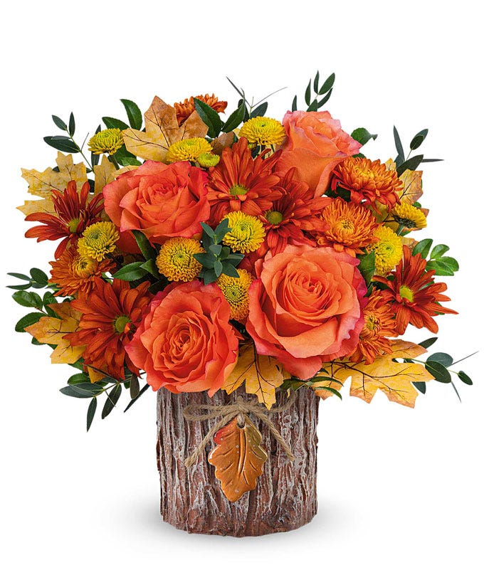 Fall floral arrangement in a rustic wooden vase adorned with a leaf accent, featuring orange roses, bronze daisy spray mums, yellow button spray mums, and seasonal greenery.