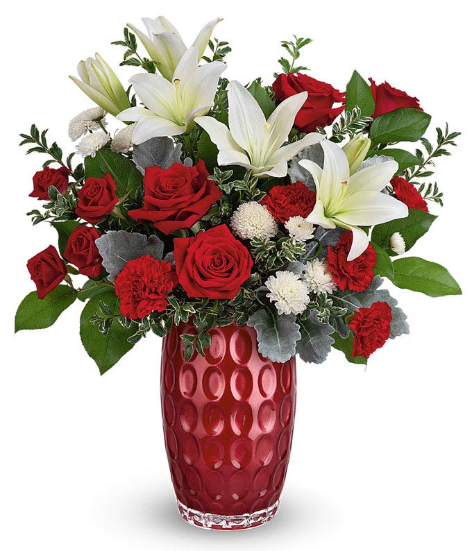 A bouquet of red roses, white asiatic lilies, mini red carnations, and spray  mums in a tall red glass vase with oval details 