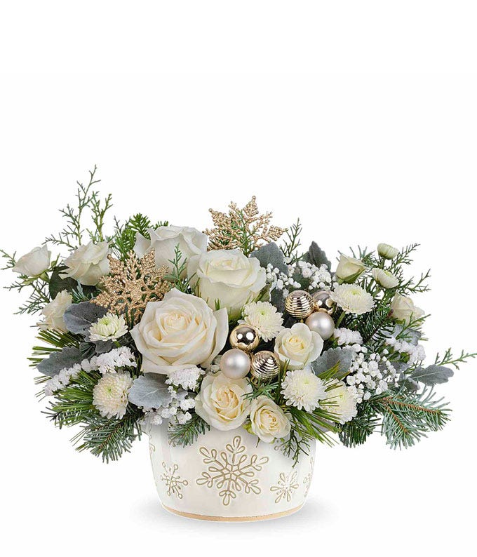 Elegant floral arrangement with white roses, carnations, cushion spray chrysanthemums, silver ornaments, frosted pinecones, snowflake accents, and evergreens in a white container with gold snowflakes.